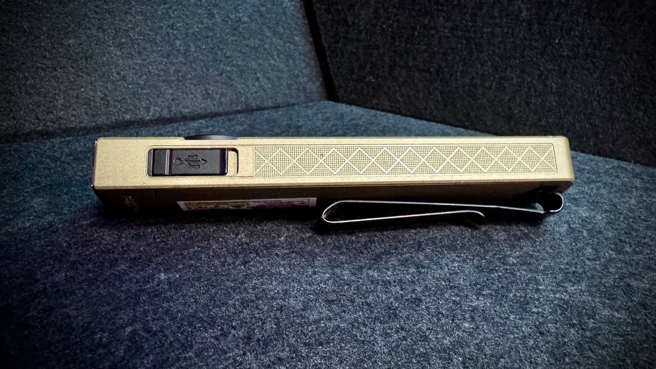 Side profile of the Olight ArkPro Ultra with the rubber flap of the USB-C charging port flipped open, showing the USB-C symbol, alongside the crosshatch knurling and the black pocket clip