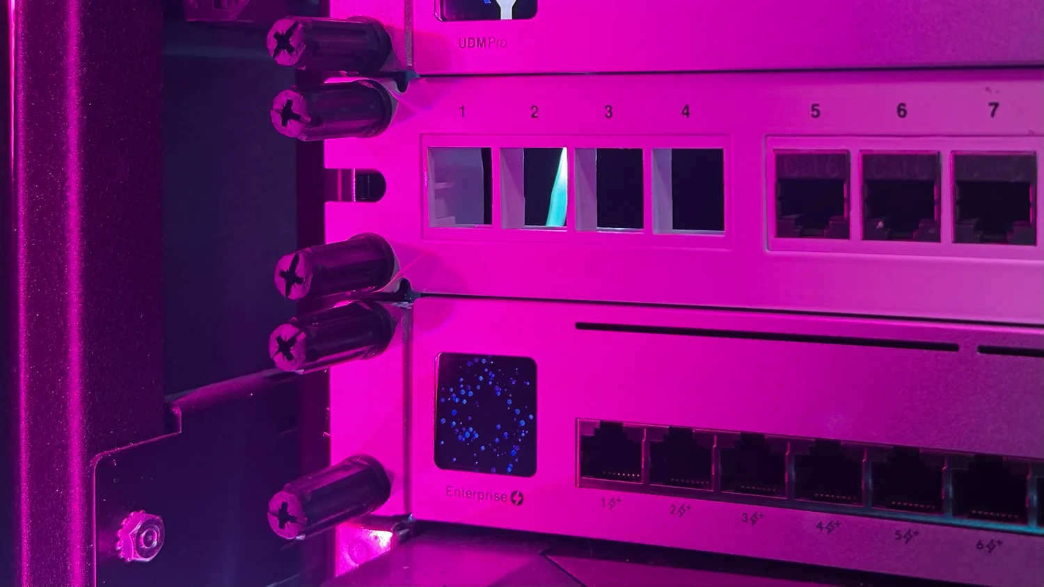 Black Rackstuds lock nuts securing a Ubiquiti UDM Pro, a patch panel and a UniFi Enterprise switch in a rack, lit in magenta, with the screwdriver slots visible on each nut